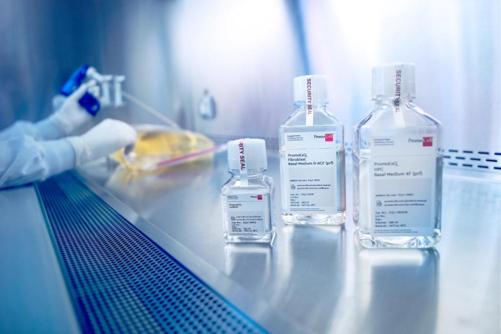 A clean bench in a laboratory containing Excipient GMP-grade media in bottles and a high-volume media bag. A person in a lab coat is working at the bench, with only their gloved hands visible as they handle materials inside.